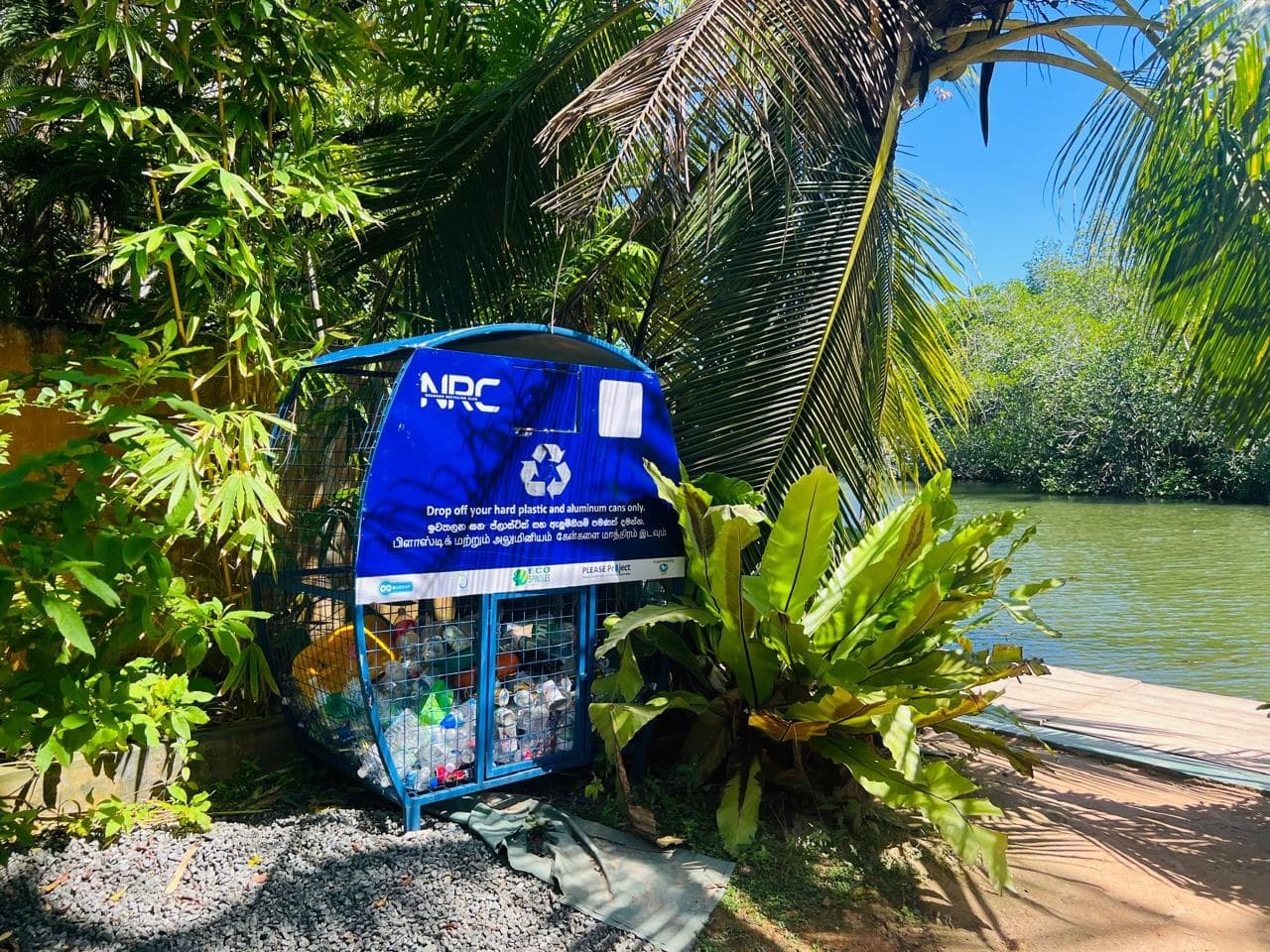 An NRC riverside drop-off bin filled with collected bottles and cans beside the waterway.